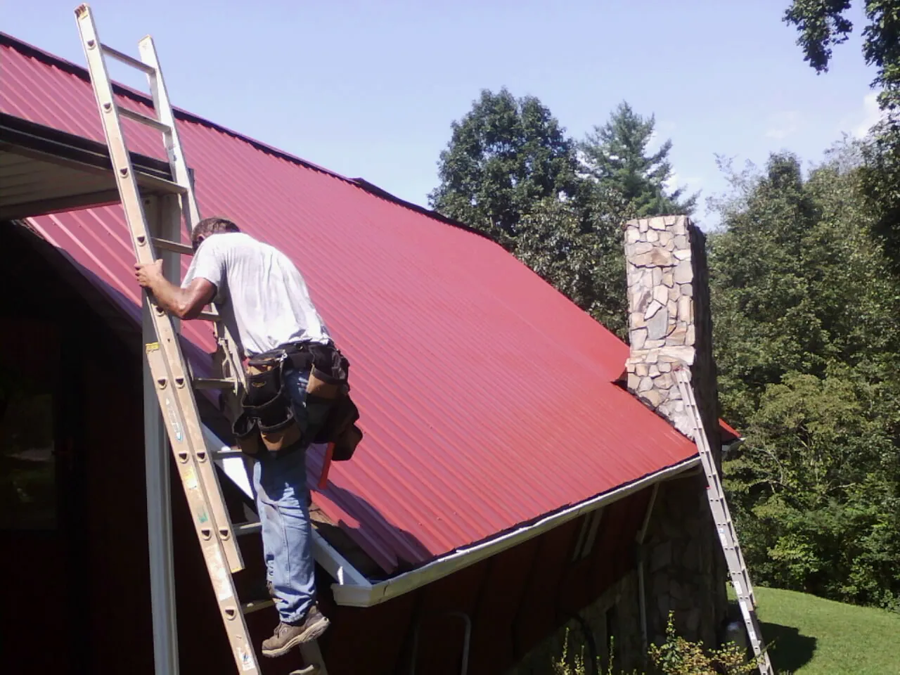 Roofer on a ladder beside a red metal roof and stone chimney — Soffit & Fascia Repair by Roofing Brothers in Rosemount